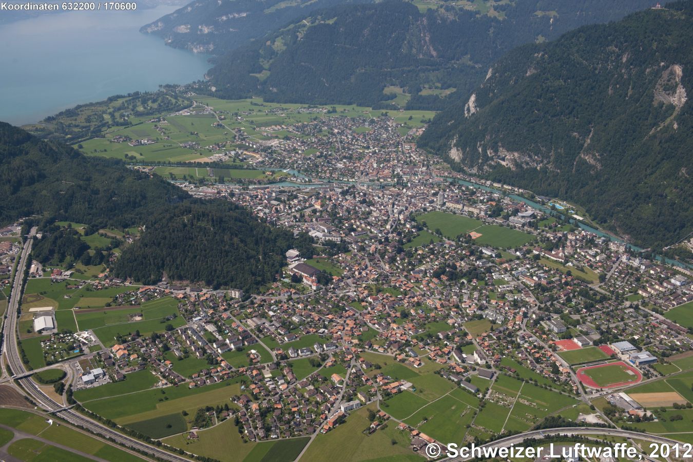 Interlaken - West (Position: 2'632'003.95, 1'169'993.49). Bildmitte: Aare-Schifffahrtskanal in den Thunersee. Im Vordergrund Autobahn A8. Bewaldeter Hügel in der rechten Mitte: Chlyne Ruuge, davor im E: Eissportzentrum Jungfrau. Rechts unten im Bild: Sportanlagen und Gymnasium. Seeufer 'Moos'.